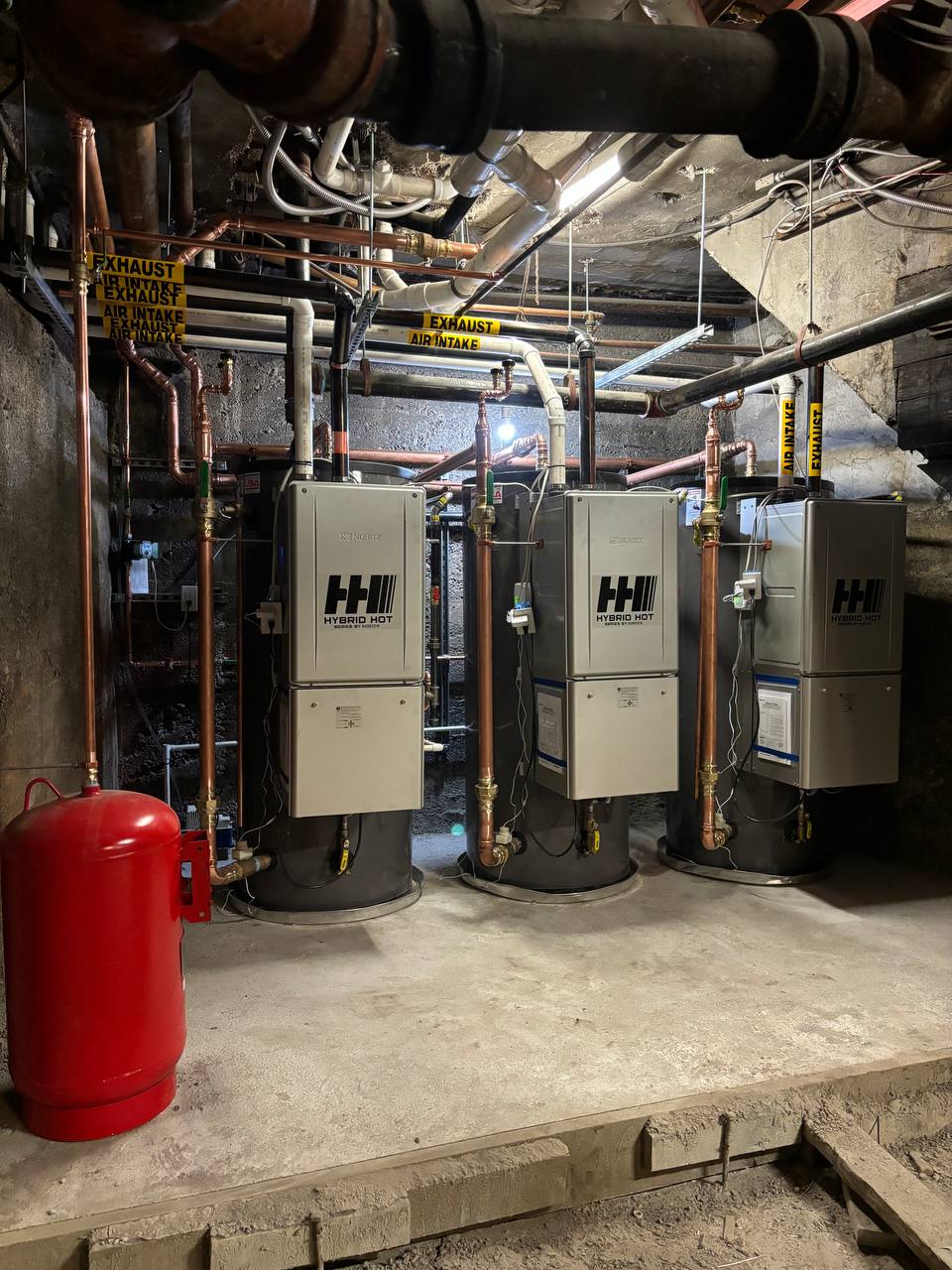 Three tankless water heaters with copper piping and red expansion tank in mechanical room